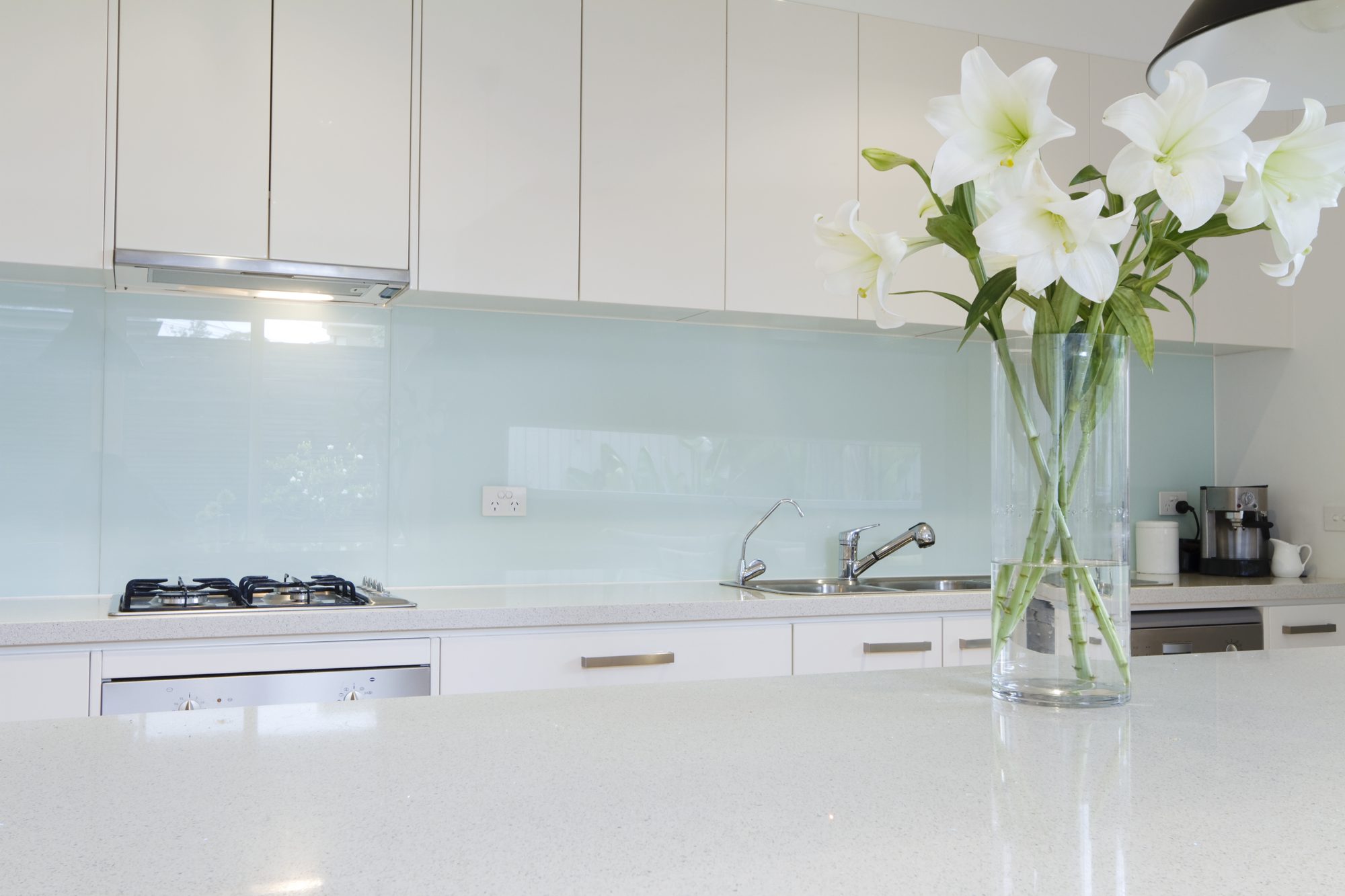 Modern white kitchen with a pale blue-green glass splashback and a vase of white lilies on the benchtop